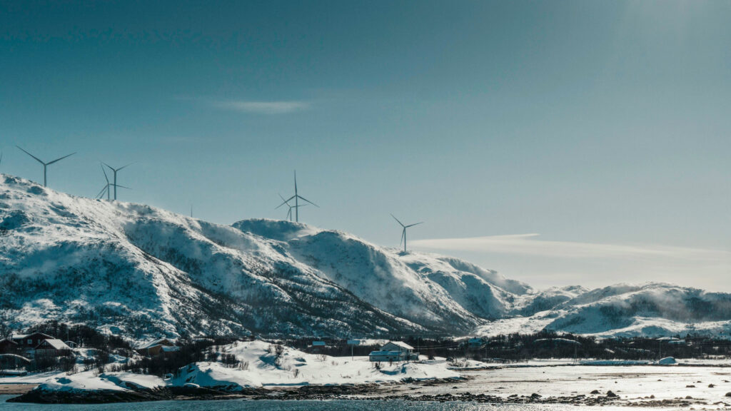 Landskap med høye fjell og snø. På toppen av fjellene er det en vindmøllepark. Sola skinner, det er småhusbebyggelse i front.