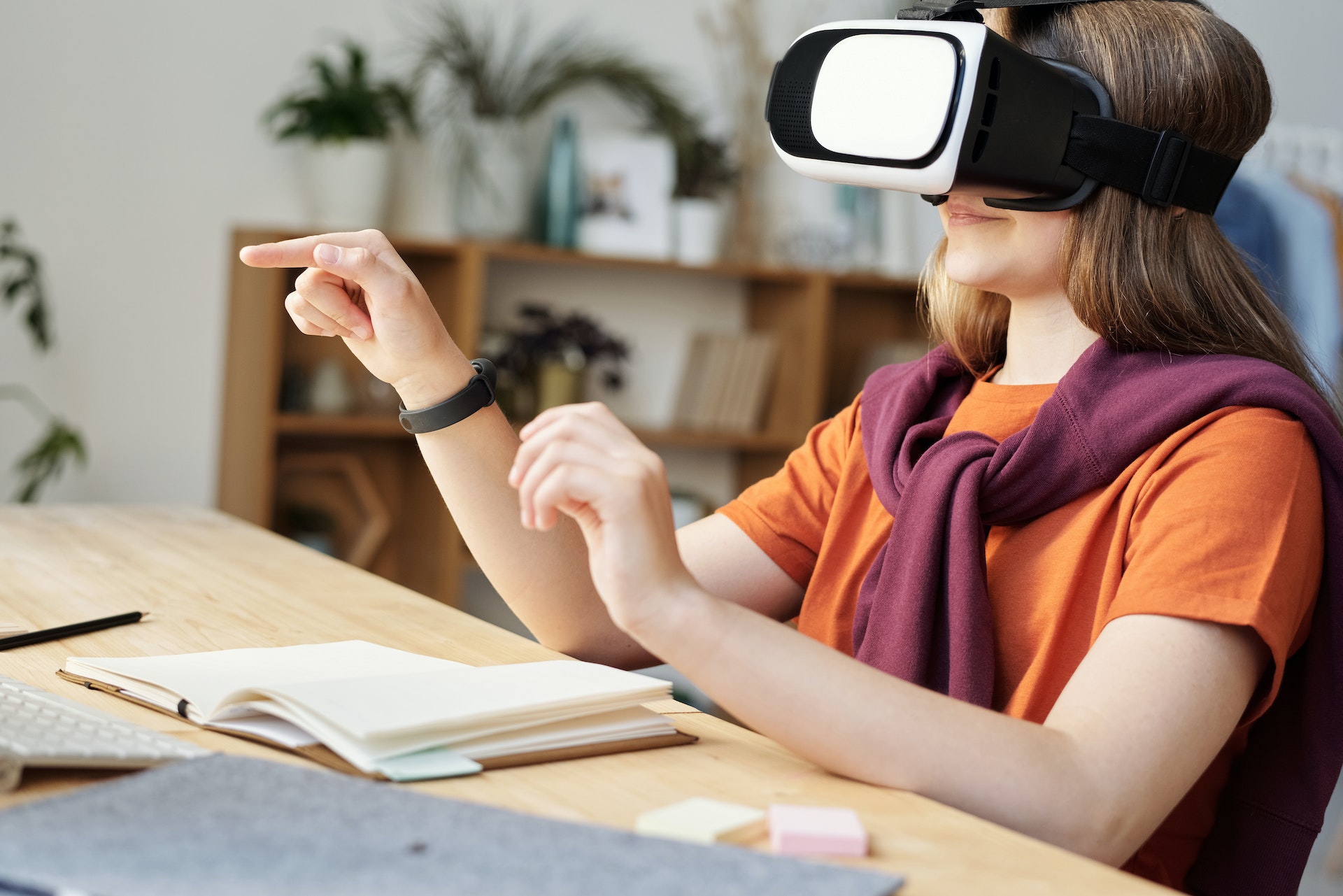 BIldet viser en jente med VR-briller på hodet. Hun sitter ved en pult, smiler og peker i lufta. Jenta har på seg en oransje t-skjorte og en burgunder collegegenser over skuldrene.