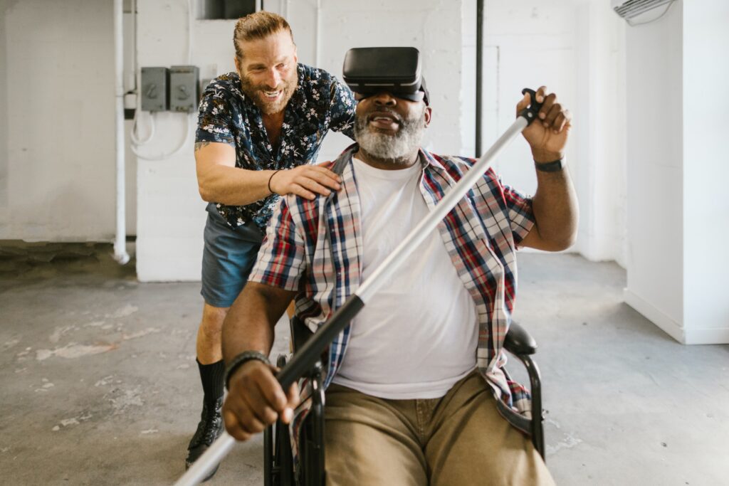 Bildet viser en mann i rullestol med VR-briller på hodet. Han holder i en padle og smiler. Det står en annen smilende mann bak og dytter rullestolen. H