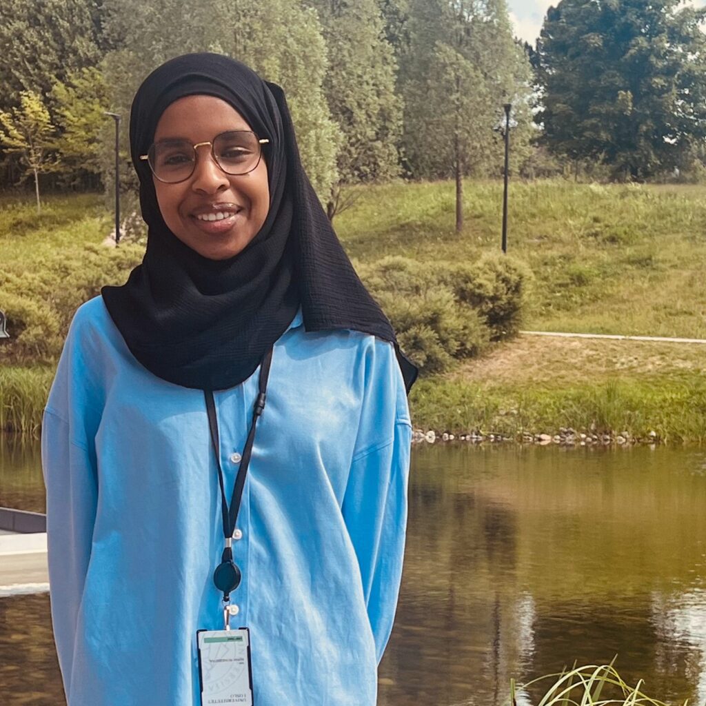 Awo står i et parkområde med vannspeil bak seg. Hun har på seg briller, blå skjorte og sort hijab. Hun ser rett mot kameraet og smiler. 