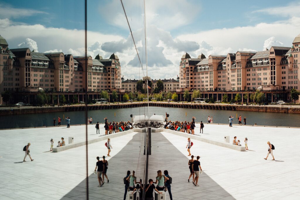 Havnelageret sett fra Operaen i Oslo. Det er klynger med mennesker som vandrer rundt Operaen og foran vannet. Det er sommer. 