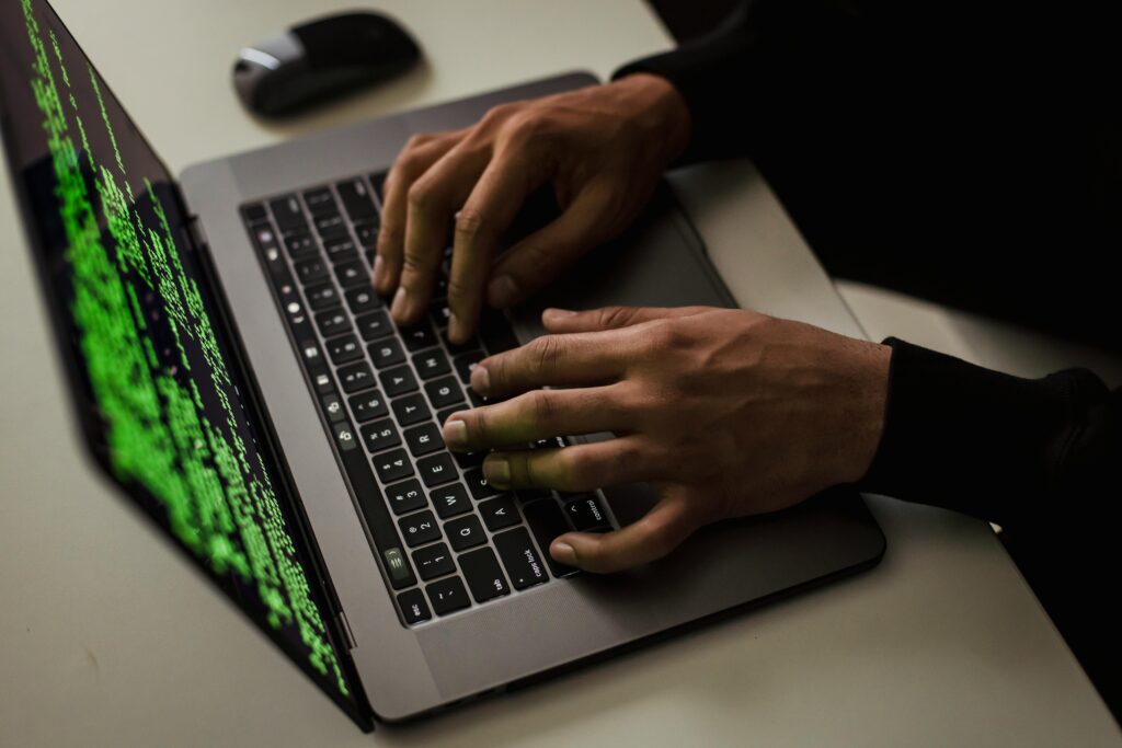 Bilde viser et menneske som sitter ved en laptop. Det eneste som er synlige er datamaskinen, med sort bakgrunn og kode på skjermen, og personens hender på tastaturet.