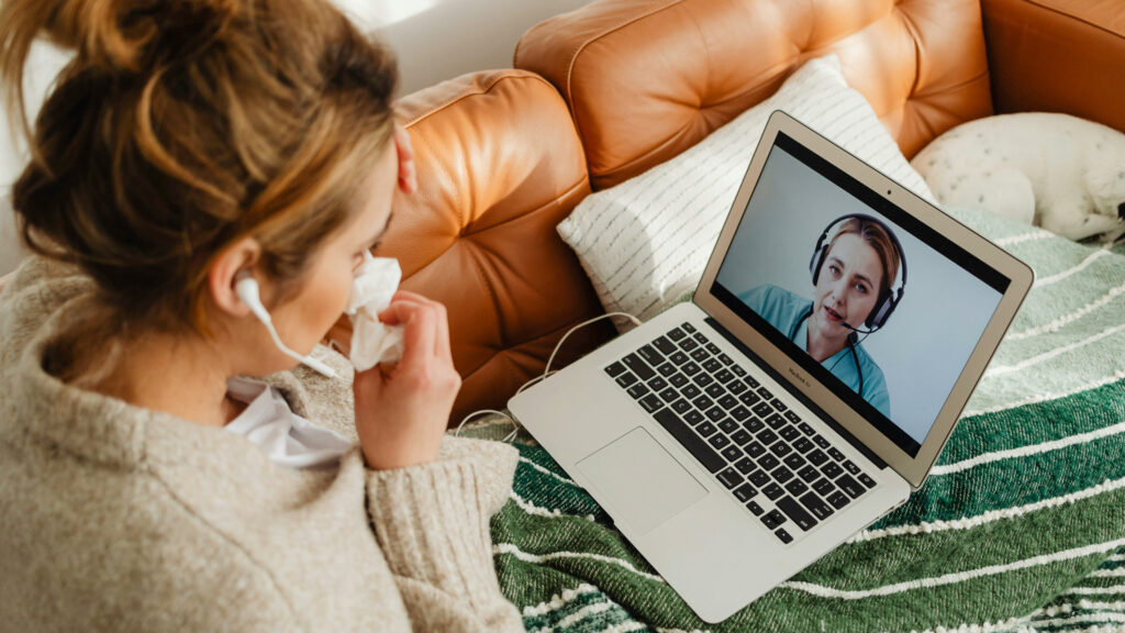 En ung kvinner sitter under et pledd på sofaen med laptop på fanget. På skjermen vises helsepersonell med headsett.
