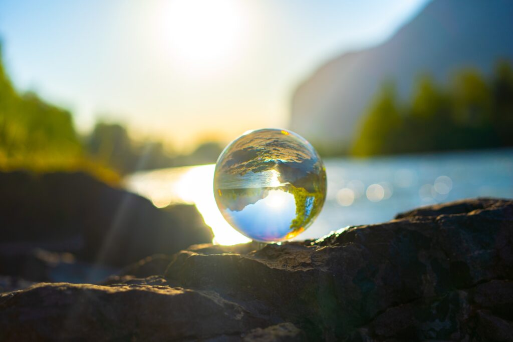 A glass ball of the Earth on a water bank. In the background blue skies, sunshine, mountains and forest can be seen.