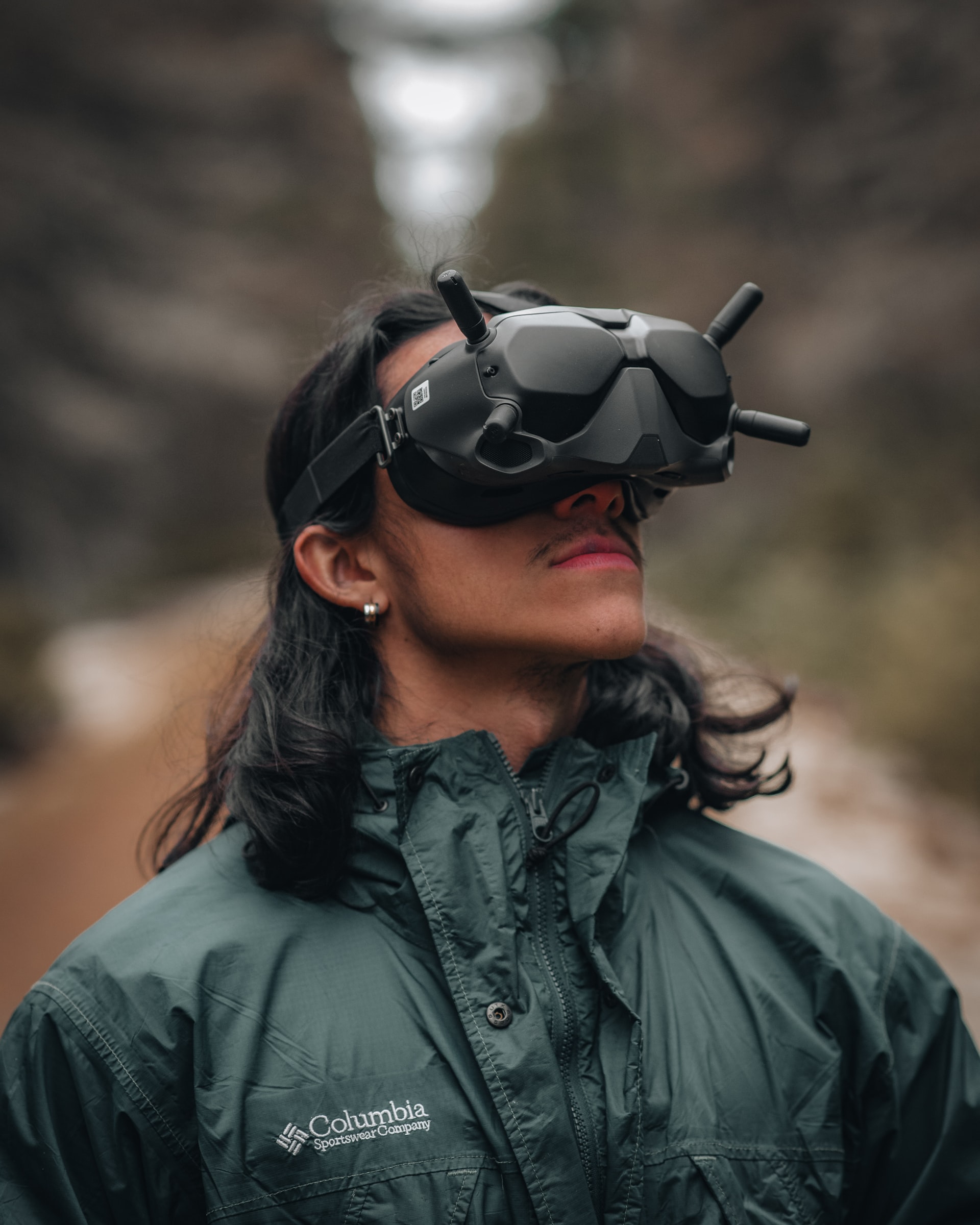 The image features a man with long dark hair and a green, Colombia anorak on. He is wearing black virtual reality goggles and looking upwards.