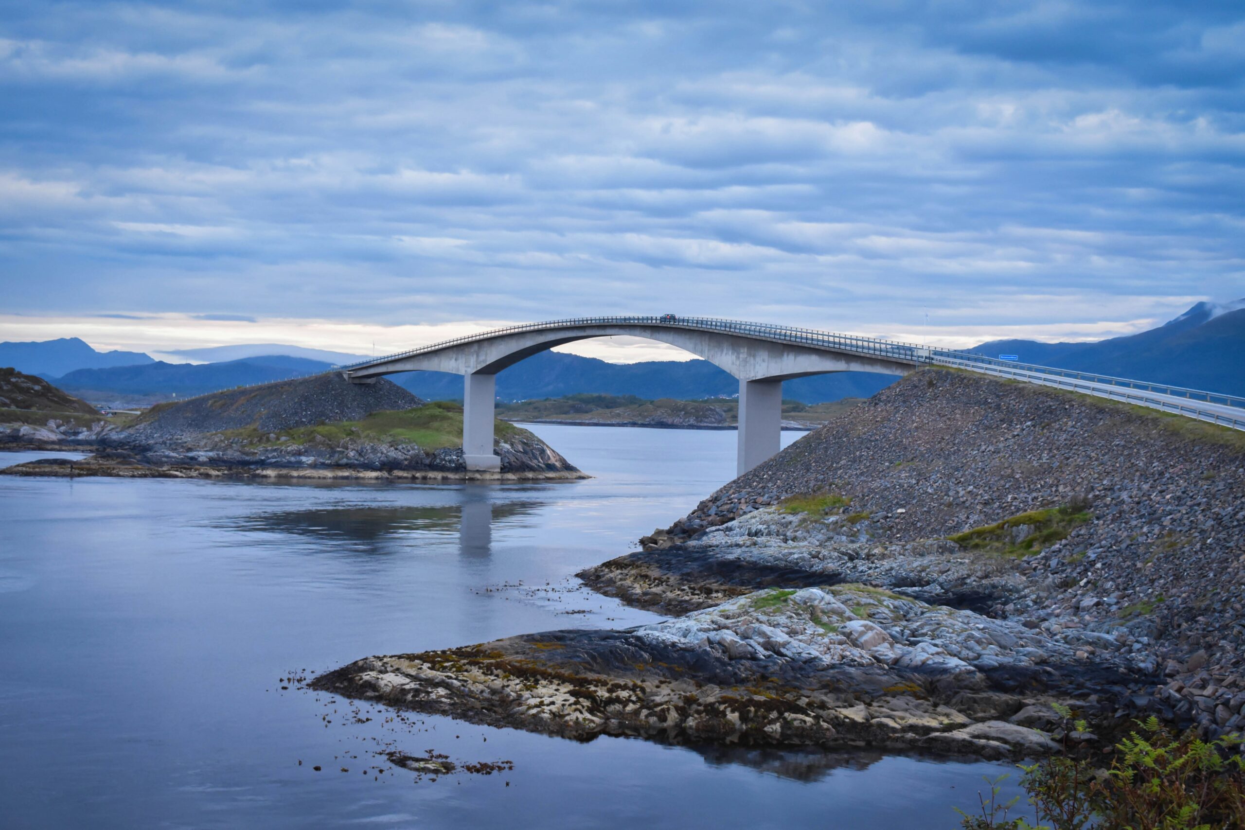 The image is a landscape shot of the bridge in Atlanterhavsvegen on the west coast of Norway. The bridge crosses a stretch of water and the land is bare and arctic. The sky is cloudy and grey.