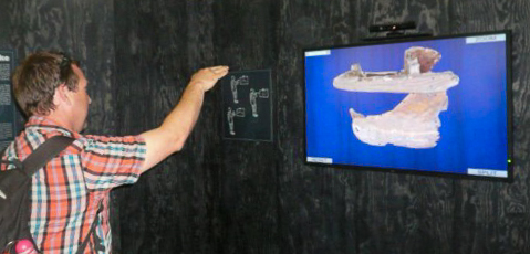 A man is seen raising his hand towards a blue screen featuring an image of a horse saddle. The image is from an interactive archaeological installation at the Viking Ship museum during the summer of 2012.