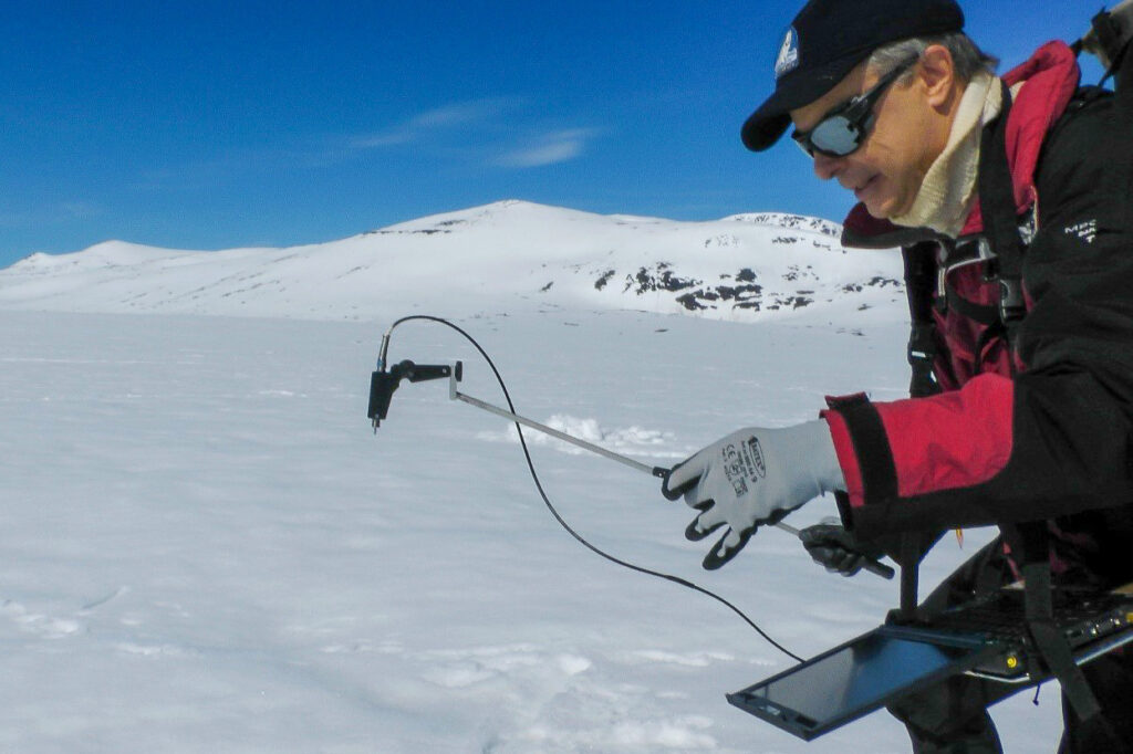 Rune is seen in an open snow landscape with monitoring equipment. He is wearing a red jacket, black cap and large sunglasses to protect from the cold and glare of the surroundings.