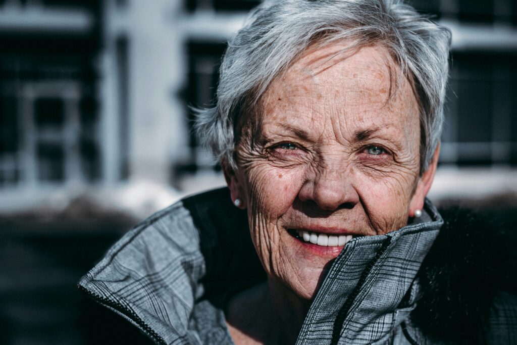 This is a close-up photo of an elderly women who is smiling towards the camera. She has short hair and a modern look. She is outside.