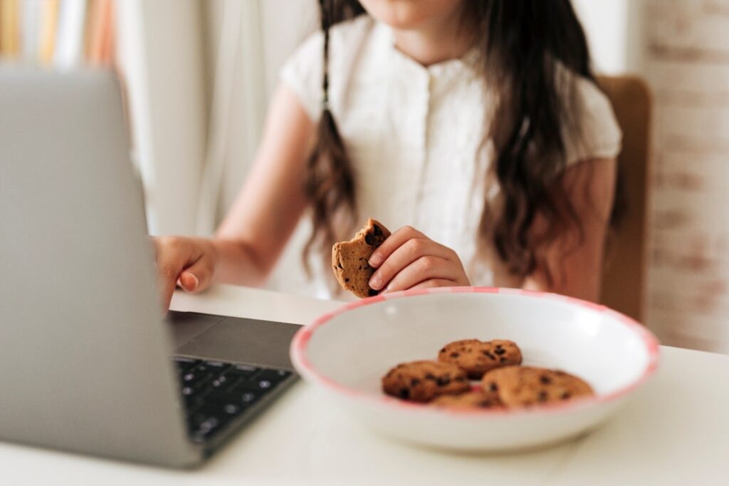 This close-up image features a young girl with partially braided dark long hair, sitting in front of a laptop. A bowl of chocolate cookies is positioned next to the laptop. The image is focused on her hands, with the right hand interacting with the laptop keyboard, and the left hand holding a cookie with a bite taken out of it. The upper part of the girl's head is not visible as it is outside the frame of the picture.