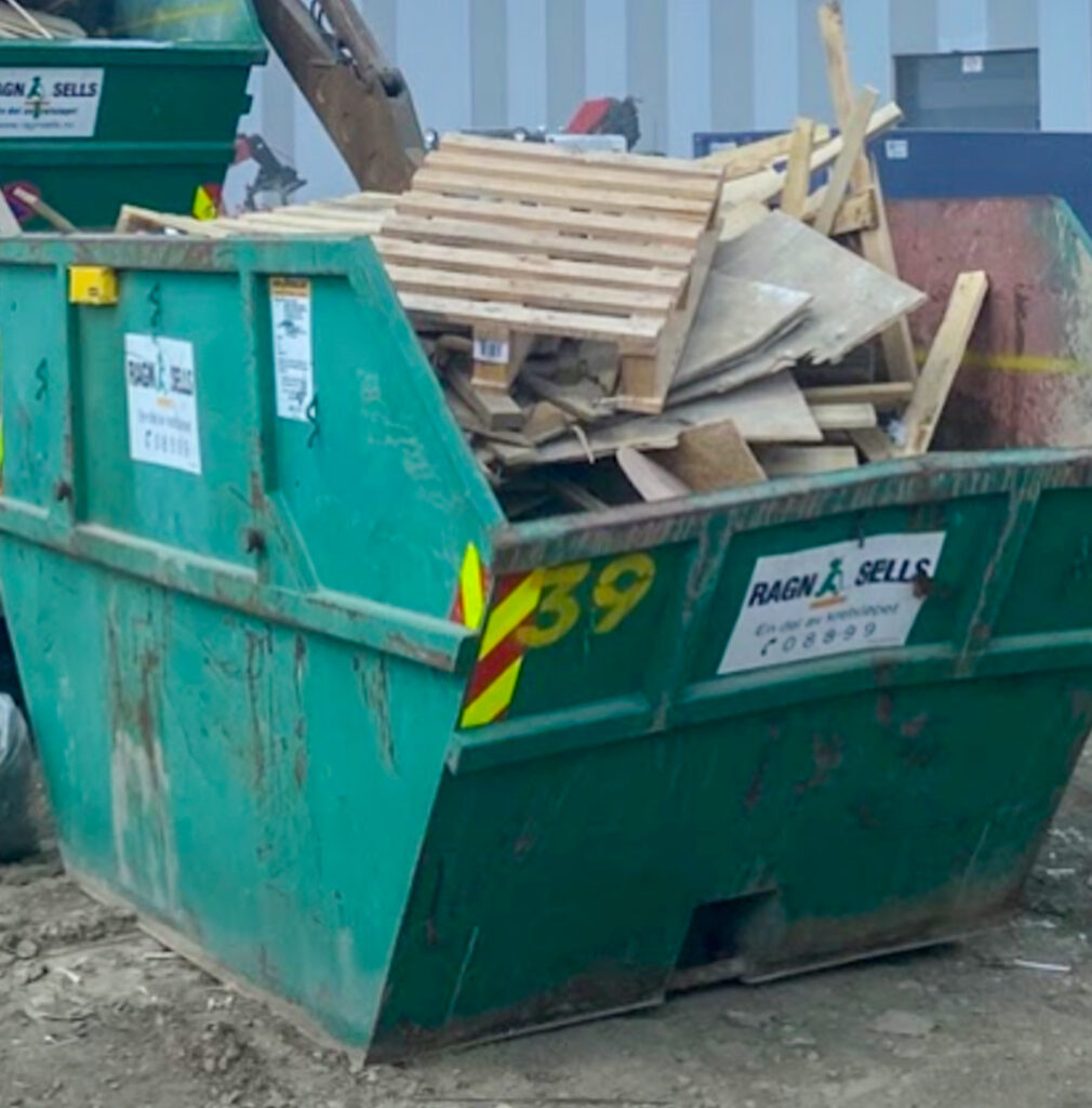 The image shows a green, open industrial waster container (a skip) with various contents piled into it. 