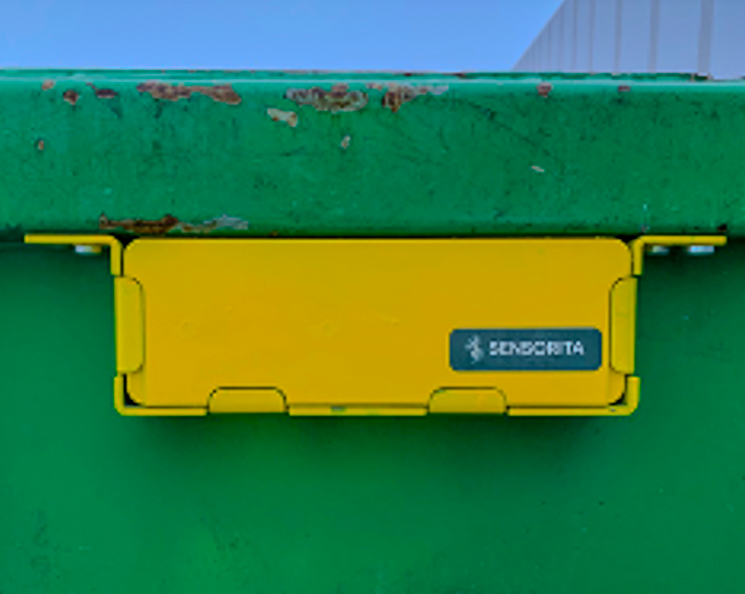 This is a close-up imahe of a sensor provided by Sensorita AS. The sensor is yellow and attached to a green waste container.