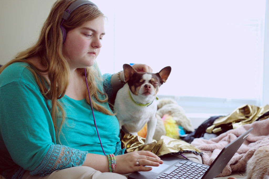 The image shows a young woman with blonde hair and a green top sitting at her laptop in a casual, home learning environment. She is patting her pet dog on the head and she looks very concentrated.