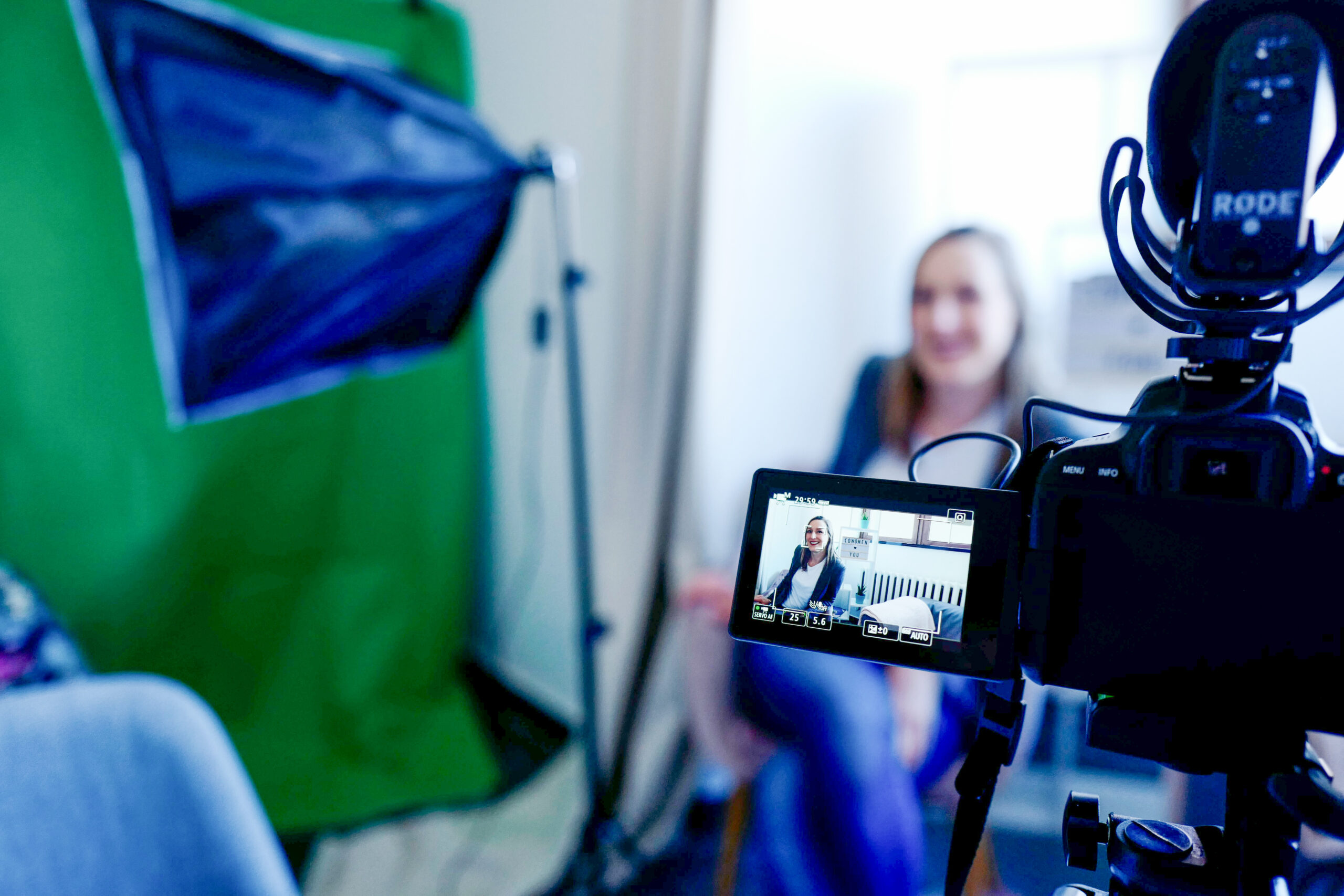 The image shows a woman in a studio setup sitting behind a mobile camera. The camera displays footage and is in focus, while the rest of the image is less sharp.