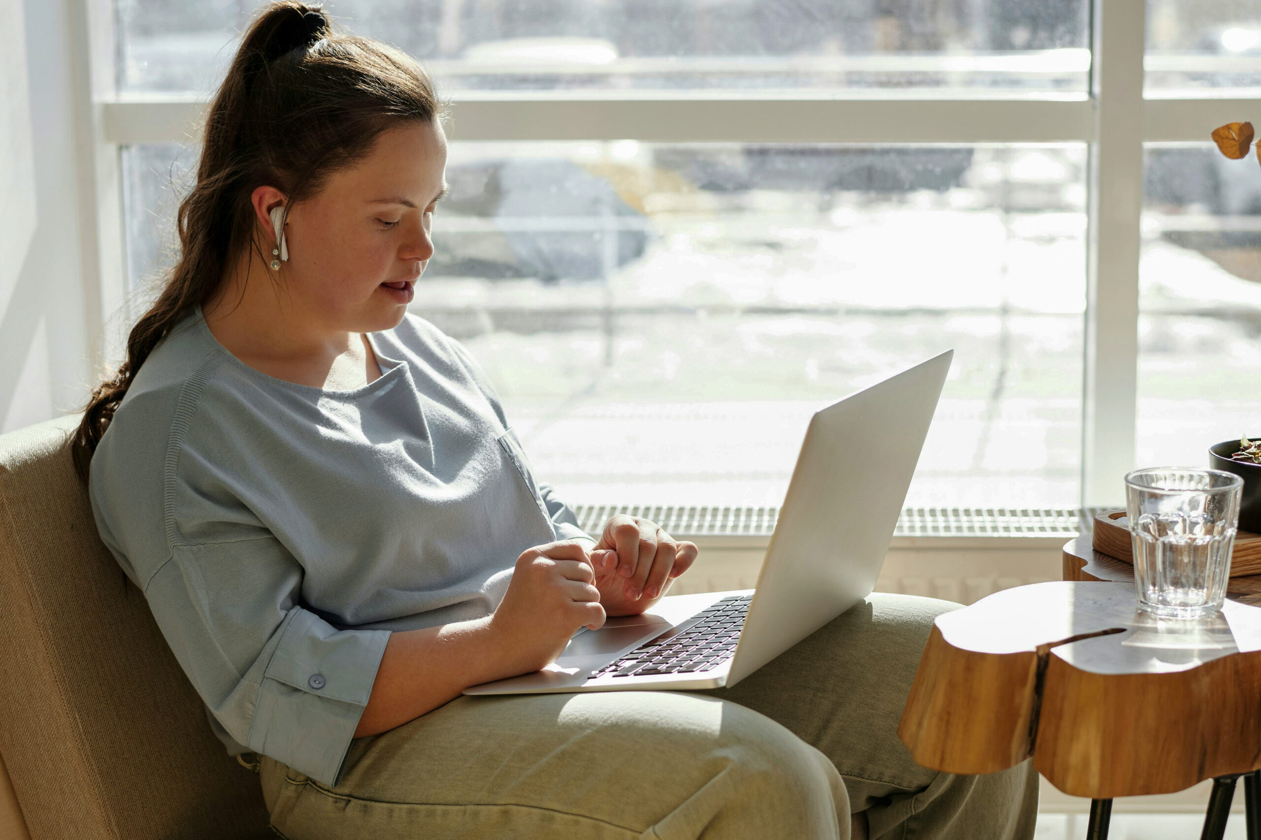 A woman with Down syndrome and dark hair is sitting in front of a bright window in a beige armchair, in front of a modern wooden coffee table. On the table, there is a glass water carafe and a vase with dried flowers. The window is lit up by the sun, and one can see a road or a parking lot with some cars that are out of focus. The woman has headphones in her ears and a laptop on her lap, and is speaking with someone on her computer.