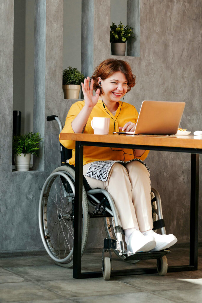 A woman with red hair ny in a wheelchair is sitting at the dining table in a very modern apartment. The floor is made of gray tiles, the wall is of red bricks, and the ceiling is painted white. Behind her is a gray concrete wall with niches housing green houseplants. The room behind her is partially in the dark, where parts of the entrance area are visible. She is communicating with someone via her laptop, greeting them by waving with her right hand. She is using headphones to both listen and speak.