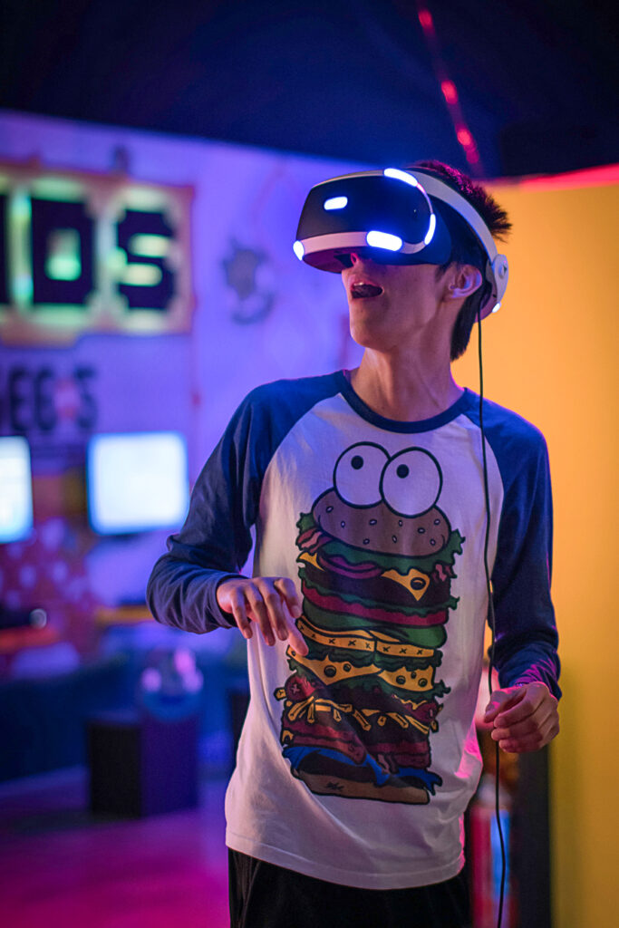 The image shows a teenage boy wearing a VR headset at a museum. The room is dark but there are bright lights in the background. The boy has dark hair and is wearing a baseball shirt with an illustrated hamburger on the front.