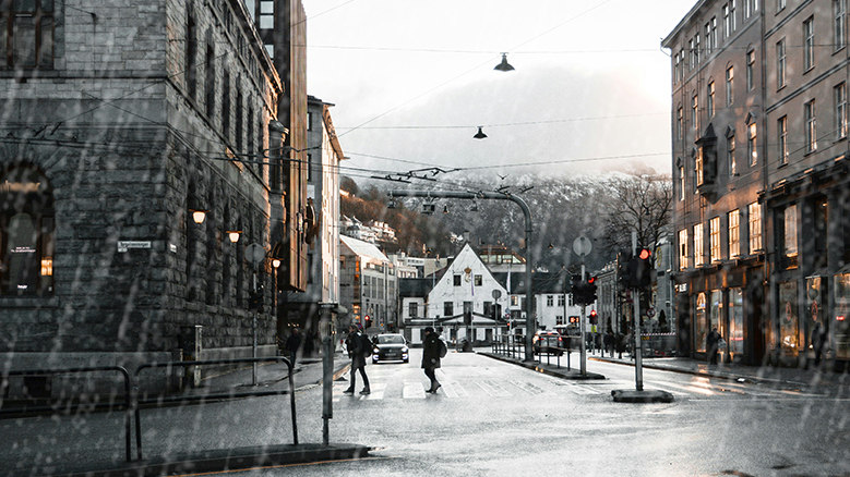 The image shows a rainy day in Bergen city centre. Buildings are on both sides of a wet street. Foggy mountains are in the background.