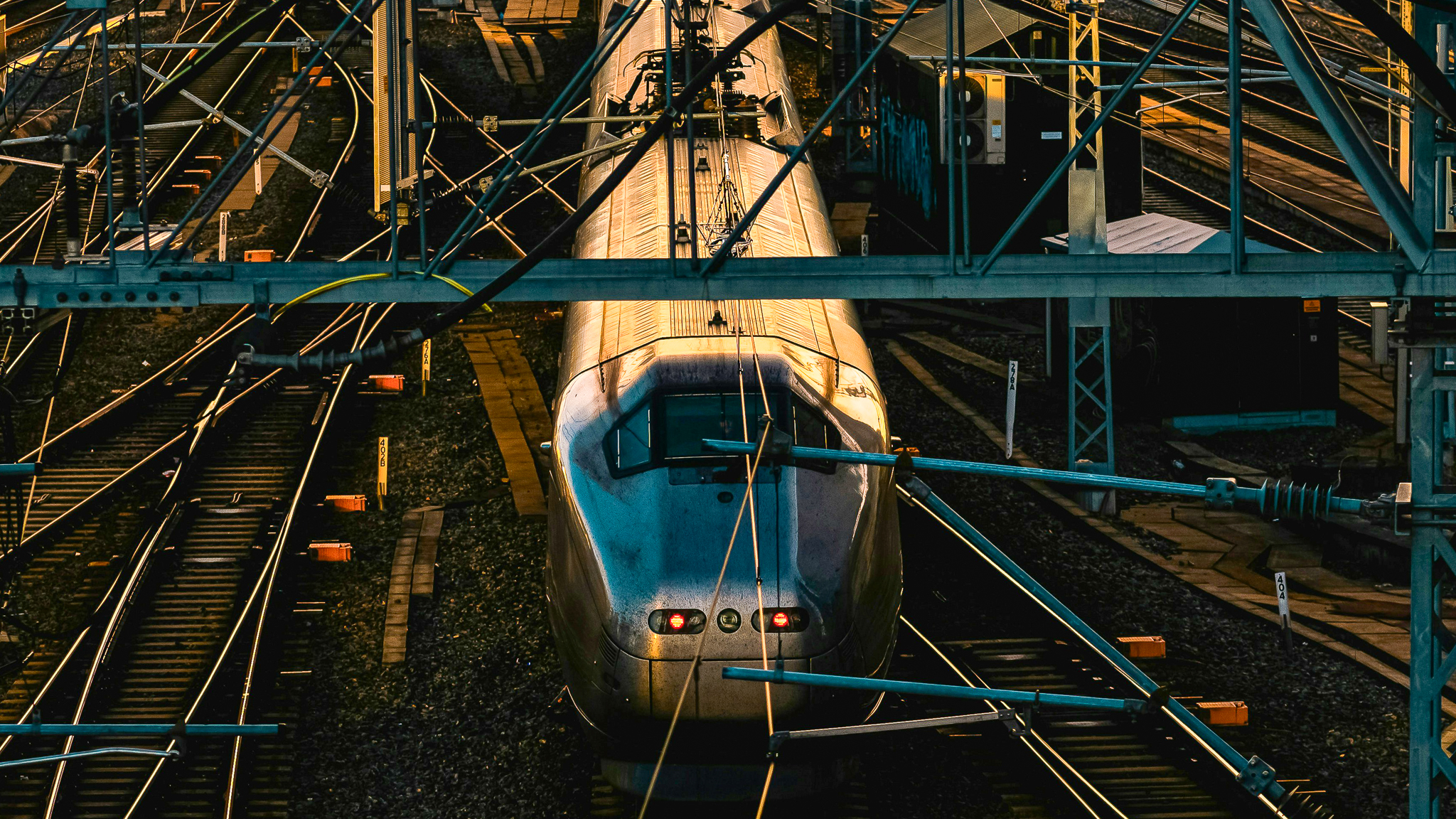 The image shows a train on the tracks with several overhead power lines and cables above it.