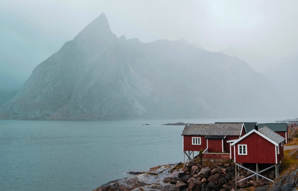 A Norwegian fishing lodge by the sea. Mountains and cloudy skies seen in the background.
