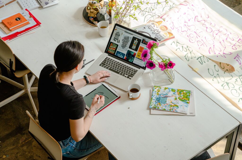Creative space. Woman sat at white desk working on a tablet in front of her laptop. Surrounded by sheets of calligraphy and magazines.